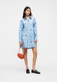 Γυναίκα όρθια, φοράει μπλε φόρεμα-πουκάμισο με λουλούδια και κουμπιά, ροζ κάλτσες, μαύρα μοκασίνια και κρατάει μια μικρή κόκκινη τσάντα στο πλάι της.