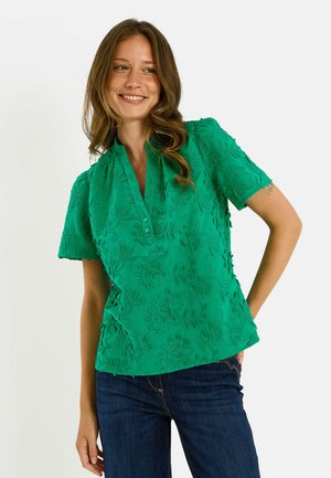 Mujer sonriente con cabello largo que lleva una blusa verde texturizada de manga corta y vaqueros azul oscuro, posando con una mano en la cadera.