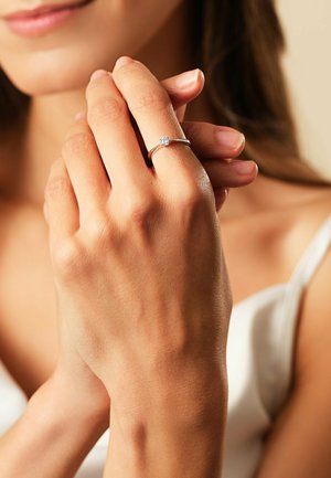 Silberner Verlobungsring mit einem quadratischen zentralen Stein, auf einem schmalen Band gesetzt, getragen an einer Hand mit natürlichen Nägeln vor einem neutralen Hintergrund.