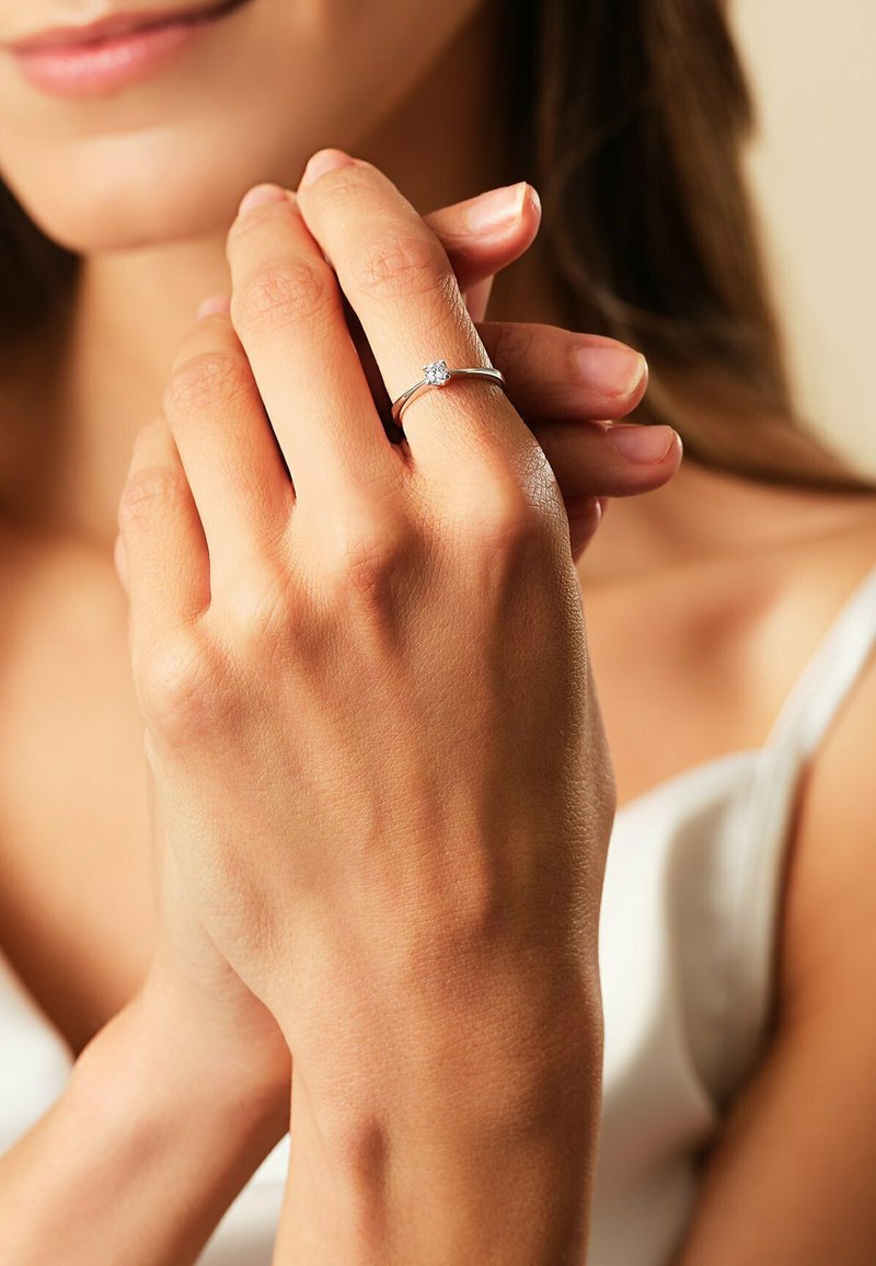 Silberner Verlobungsring mit einem quadratischen zentralen Stein, auf einem schmalen Band gesetzt, getragen an einer Hand mit natürlichen Nägeln vor einem neutralen Hintergrund.