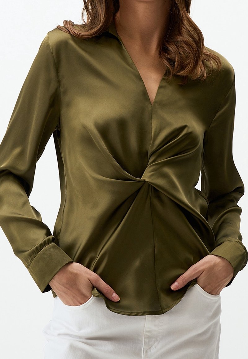 Femme portant un chemisier en satin vert olive avec un détail de nœud torsadé à la taille, associé à un pantalon blanc, les mains dans les poches.