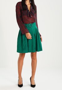 Blusa a zigzag rossa e blu con colletto arricciato abbinata a una gonna verde testurizzata con fibbia circolare e pieghe.