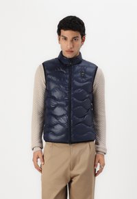 Mørkeblå puffervest med høy krage, quiltet tekstur og frontglidelås. Brukt over en lys beige, teksturert langermet skjorte og beige bukser.