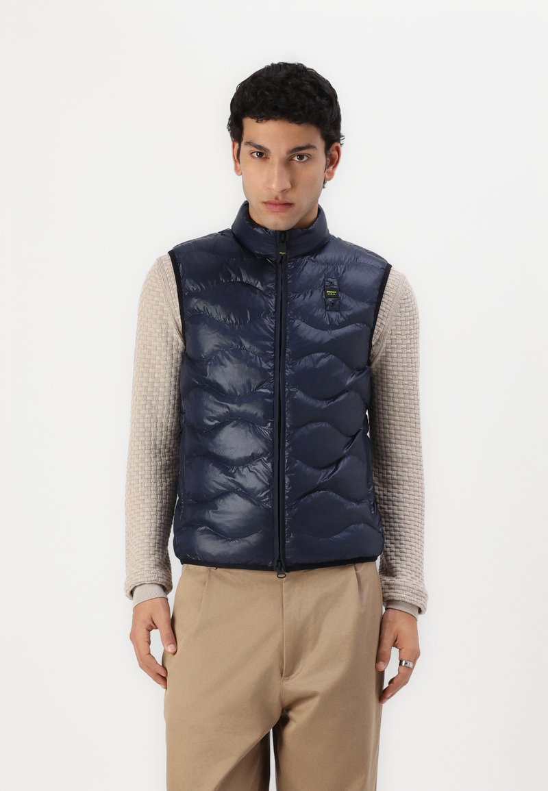 Mørkeblå puffervest med høy krage, quiltet tekstur og frontglidelås. Brukt over en lys beige, teksturert langermet skjorte og beige bukser.