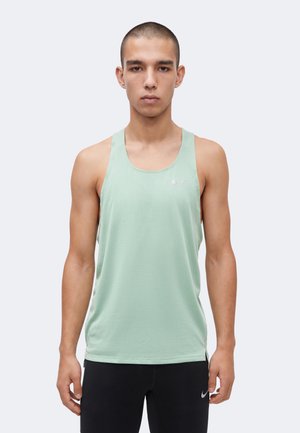 Mladý muž s krátkymi vlasmi, ktorý má na sebe svetlozelené atletické tielko a čierne nohavice, stojí na jednoduchom pozadí.