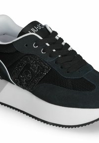 Zapatilla negra con parte superior de malla texturizada, acentos de ante y un parche de logo brillante. Cuenta con cordones negros, entresuela blanca y suela de goma.