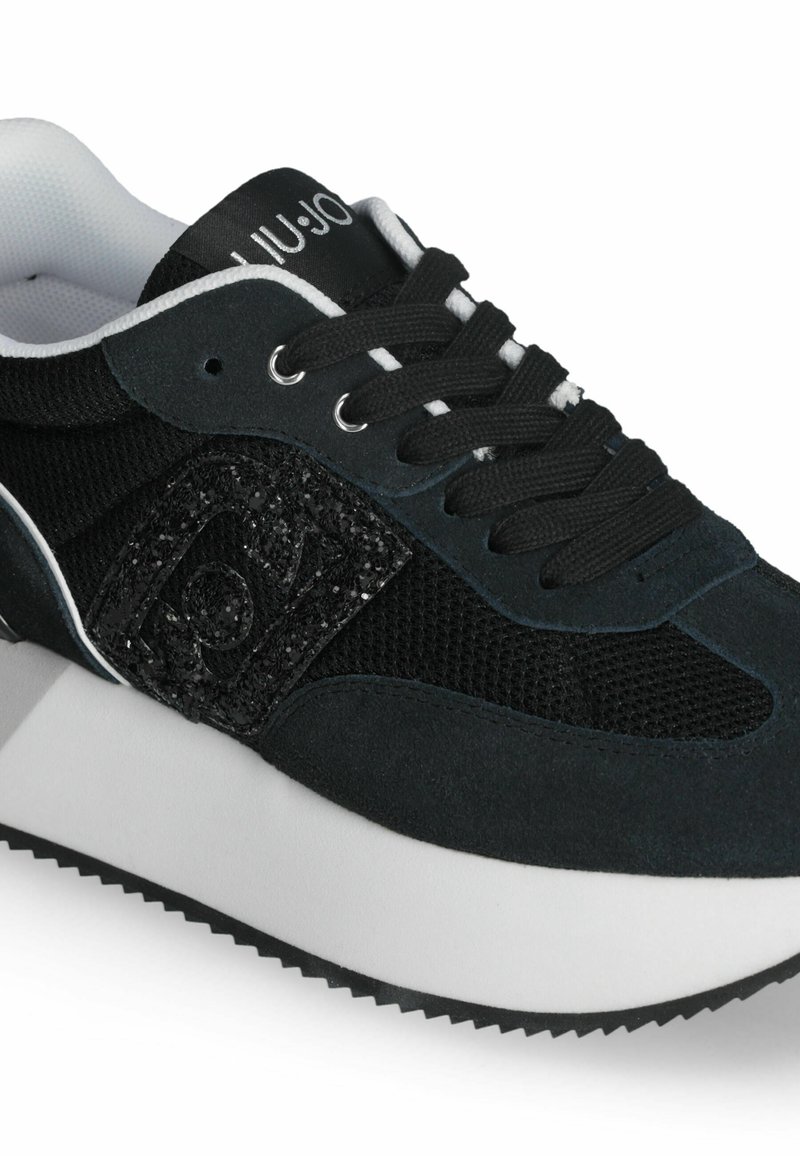 Sneaker nera con parte superiore in rete testurizzata, dettagli in suede e un'etichetta con logo luccicante. Presenta lacci neri, suola intermedia bianca e suola in gomma.