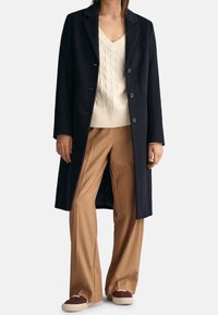 Marineblauwe wollen blend jas met een getailleerde pasvorm, enkel geknoopte sluiting, drie knopen en klepjes, gedragen met beige wijde broek.