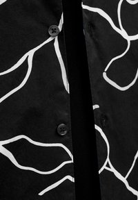 Camisa de tela negra con un patrón abstracto de líneas blancas, cierre de botones y costuras negras visibles, que presenta una textura suave.