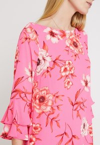 Nainen, jolla on yllään kirkkaan vaaleanpunainen paita, jossa on punaisia ja valkoisia kukkakuosisia yksityiskohtia ja kellottavat hihat, näkyy olkapäistä vyötärölle saakka.