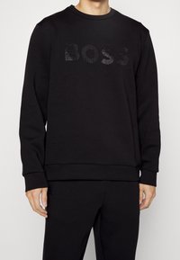 Man som bär en svart långärmad sweatshirt med en texturerad "BOSS"-logotyp över bröstet, tillsammans med matchande svarta byxor, med avslappnade händer vid sidorna.