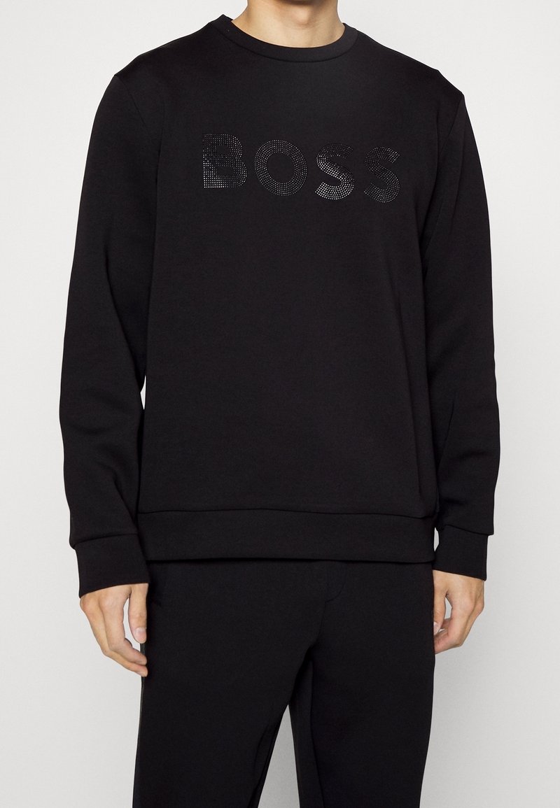 Man som bär en svart långärmad sweatshirt med en texturerad "BOSS"-logotyp över bröstet, tillsammans med matchande svarta byxor, med avslappnade händer vid sidorna.