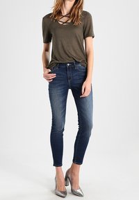 Oliivinvihreä lyhythihainen paita ristikkäisellä kaula-aukolla, yhdistetty tummansinisien slim fit -farkkujen ja metallinhohtoisilla hopeisilla korkokengillä.
