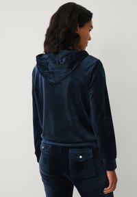 Sudadera de terciopelo azul marino con dobladillo y puños acanalados. Cuenta con una capucha forrada con detalles de volantes y una textura suave.