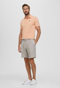 Różowa koszulka polo z przednią kieszenią, połączona z jasnoszarymi szortami i białymi sneakersami. Model stoi w pozycji wyprostowanej na neutralnym tle.