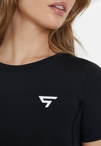 Čierny športový top vyrobený z hladkého materiálu, s priliehavým strihom a bielym logom na ľavej oblasti hrude. Kolo výstrih a krátke rukávy.