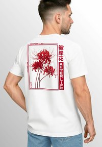 Weißes T-Shirt mit einem roten Grafikmotiv von roten Spinnenlilien, umrahmter Text mit den Worten "Rote Spinnenlilien" und "Jenseits" in fetter roter Schrift.