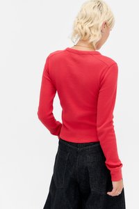 Person med kort blond hår iført en teksturert rød langermet topp og høyhelte svarte bukser, sett fra bakgrunnen mot en hvit bakgrunn.