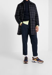 Homme portant un manteau noir à carreaux, un pantalon bleu marine, des baskets épaisses marron, bleu marine et jaune, avec des chaussettes blanches et des poignets de chemise vert néon visibles.