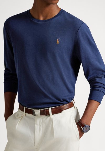 Tengerészkék hosszú ujjú ing puha anyagból, a mellkasán kis hímzett logóval, krémszínű nadrággal és barna övvel párosítva.