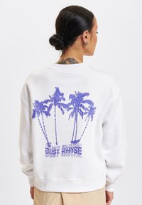 Weißes Sweatshirt mit lila Palmen-Motiv und dem Text "JUST RINSE" auf dem Rücken. Verfügt über gerippte Bündchen und Saum sowie eine weiche Stofftextur.