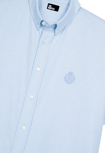 Camisa de botones azul claro hecha de una tela de textura suave, con un cuello clásico y un emblema bordado en el lado izquierdo del pecho.