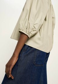 Dunkelhäutige Person trägt eine beige Bluse mit Puffärmeln und einen dunkelblauen Jeansrock, die Hand entspannt an der Seite vor einem einfarbigen Hintergrund.