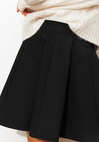 Jupe plissée noire en tissu texturé et extensible. Dotée d'une taille haute et d'une silhouette évasée, avec des coutures soignées et une longueur au-dessus du genou.