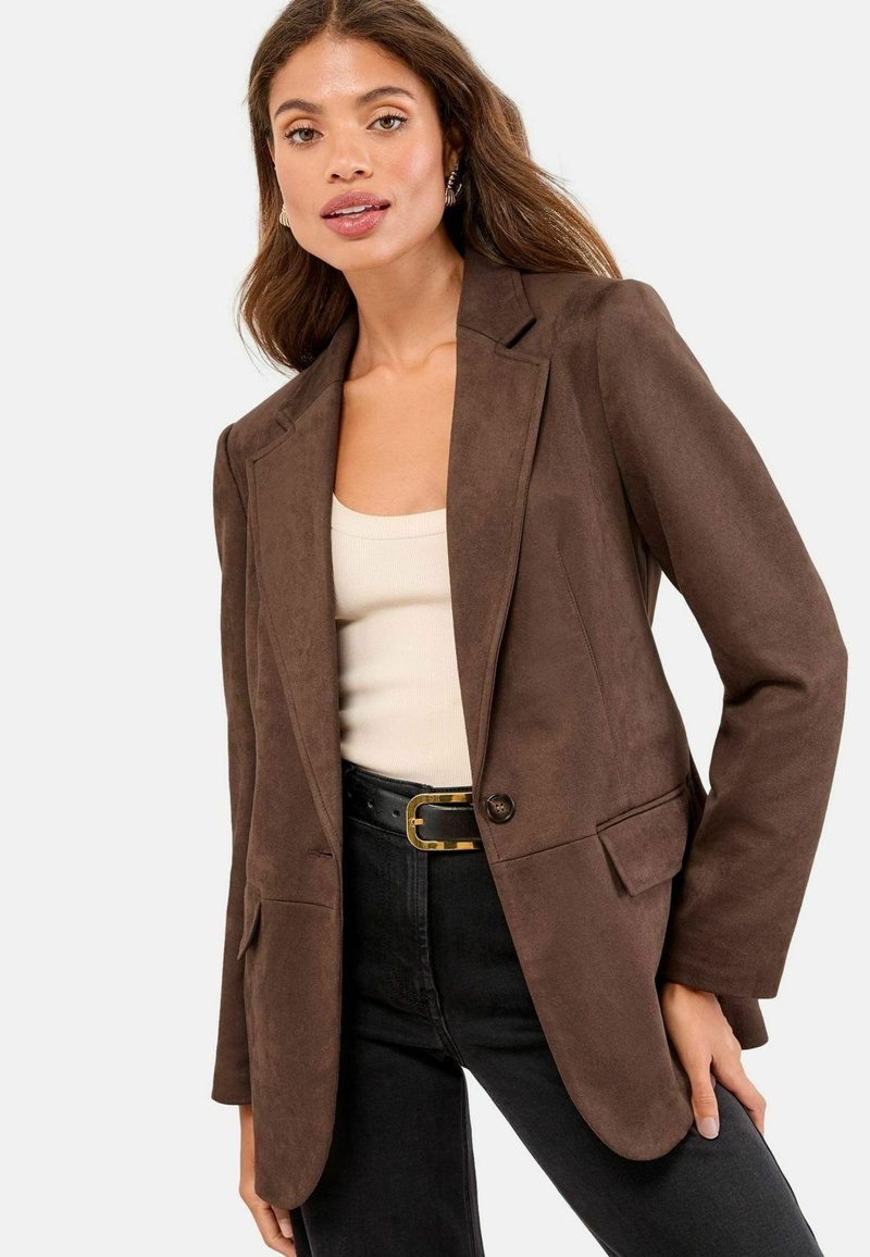 Hnedý blazer z imitácie semišu s klopovými goliermi, jedným gombíkom a dvoma prednými vreckami. Skombinovaný s krémovým topom a čiernymi džínsami.