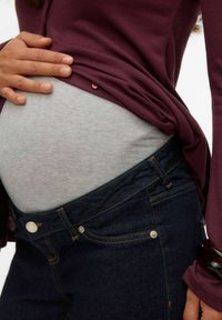 Graviditetsklädsel med en mörk lila långärmad tröja, grå underkläder och mörka denimjeans med gyllene sömmar och metallaccenter.