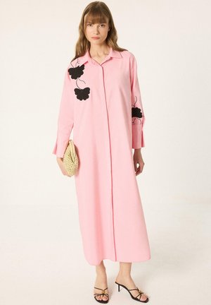 Femme portant une longue robe rose avec des motifs floraux noirs sur les manches et la poitrine, tenant une pochette tissée, portant des sandales noires à talons.