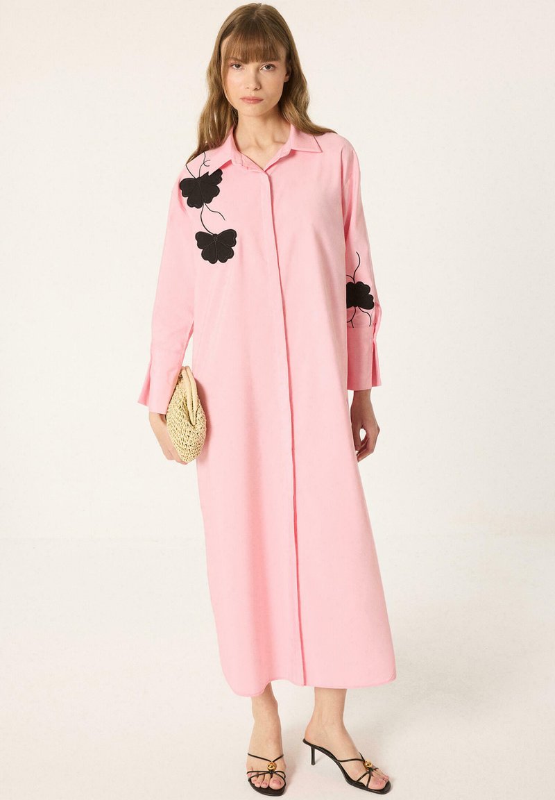Femme portant une longue robe rose avec des motifs floraux noirs sur les manches et la poitrine, tenant une pochette tissée, portant des sandales noires à talons.