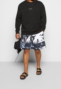 Sweatshirt noir avec le logo blanc de Calvin Klein, shorts de bain noirs et blancs à motifs, sandales noires, portant un sac noir.