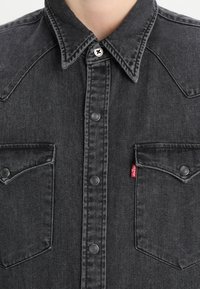 Camicia in denim nero con colletto a punta, chiusura con bottoni a pressione, due tasche sul petto e un piccolo dettaglio con linguetta rossa sul lato sinistro.