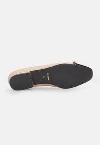 Sole di scarpa con tacco color beige, con superficie liscia, dotata di una base in gomma nera, marchio dorato e forma arrotondata, leggermente appuntita.