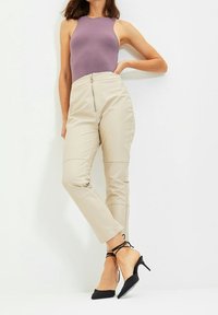 Vrouw in een mouwloze lavendelkleurige top, hoge beige broek met een ritssluiting aan de voorkant, en zwarte puntige hakken met enkelbandjes, staand tegen een witte muur.