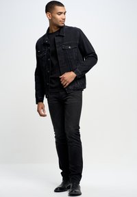 Giacca di denim nera con tasche frontali, abbinata a una camicia nera e jeans scuri. Tessuto liscio con una silhouette aderente e dettagli minimali.