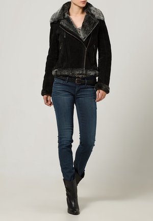 Veste en daim noire avec un col et des poignets en fourrure grise, dotée d'une fermeture éclair asymétrique et de poches latérales. Portée avec un jean bleu et des bottes noires.