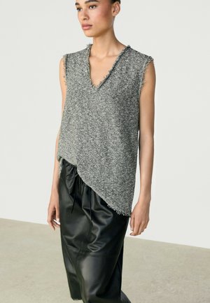 Femme portant un haut gris en maille sans manches avec des bords effilochés et un pantalon en cuir noir, debout sur un fond clair uni.
