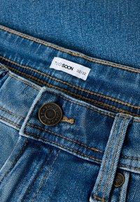 Sinised teksad, kus vöökoht näitab metallist nuppu, millel on kiri "Two Soon Jeans", ning valget suurusega 32/32 rõivilabelit.