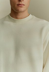 Felpa beige chiaro con un motivo a diamante testurizzato, colletto a girocollo a costine e tessuto morbido. Inquadratura ravvicinata che mette in evidenza la sua forma e il design.