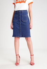 Denim A-line skirt with front buttons, two front pockets, raw hem, and a mid-blue wash. Worn with a white t-shirt and red sandals.