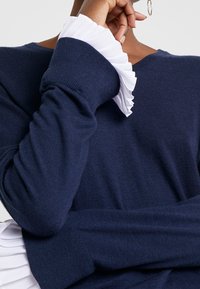 Granatowy sweter wełniany z długim rękawem, z białymi falbaniastymi mankietami. Materiał wydaje się miękki i teksturowany, z luźnym krojem.