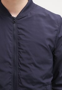 Námořnická bomber bunda se zipem vpředu, žebrovaným límcem a mírně nadýchanou texturou. Má hladkou látku a minimální šití.