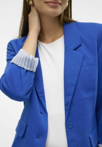 Niebieska marynarka z usztywnionymi ramionami, zapinana na jeden guzik, z haftowaną kieszenią na piersi. Biały top z widocznymi mankietami w jasnoniebieskie paski.