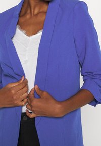 Osoba v obleku s módním fialovým sako přes bílé tričko, který si upravuje knoflíky oběma rukama a předvádí upravený a moderní vzhled.