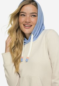 Sweatshirt med en cremefarvet krop, blå hætte og kontrasterende snore. Har en forlommeklud og struktureret stof.