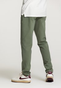 Pantaloni jogger verzi din bumbac, cu o croială conică, având o talie elastică, buzunare laterale și o textură netedă. Îmbrăcați cu adidași albi și vișinii.
