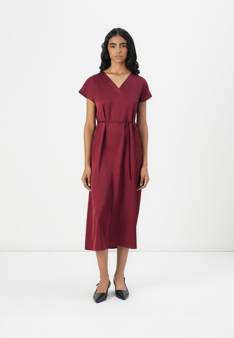 Robe midi bourgogne à manches courtes, décolleté en V et ceinture à la taille. Texture lisse et brillante. Portée avec des chaussures noires à bout pointu.