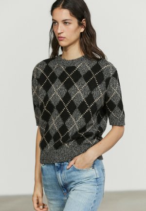 Donna che indossa un maglione argyle nero e grigio a maniche corte con jeans azzurri, in piedi con una mano in tasca.